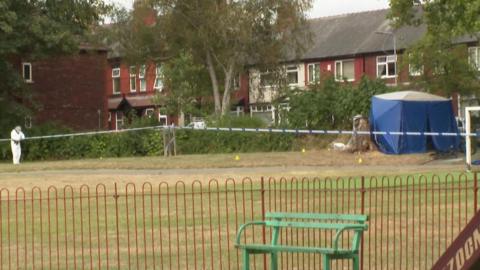 A forensic officer in white protective clothing is standing on a grassed area, with a tent near to some fencing.
