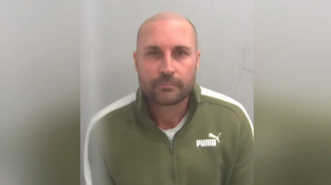 A mugshot of David Richards. He is bald with stubble and is wearing a green sip-up jumper with a Puma logo to the right.