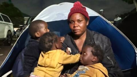 Esnat Joseph, wearing a maroon woollen bobble hat and black jacket, sitting in front of a tent with her baby triplets - two dressed in yellow hoodies and one black.