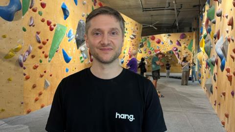 Jon Partridge standing head with a grin. He is wearing a black t-shirt with the Hang company logo. Climbing walls with his customers standing can be seen behind him.