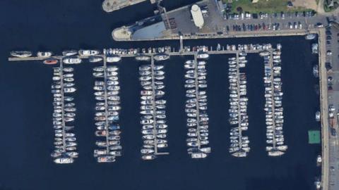 Aerial view of the marina. There are six long pontoons leading off one large attached to the harbour car park by a gangplank. Dozens of boats are tightly packed along the pontoons.