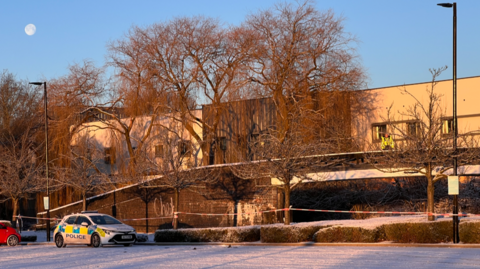 A police officer is standing on top of a bridge, with a police car parked to the side. There is red and white tape under the bridge
