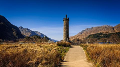 Glenfinnan Monument is a tower-like structure with a statue on top of it. The monument is surrounded by mountains and hills with Loch Shiel in the background. The scene is under a beautiful blue sky. 