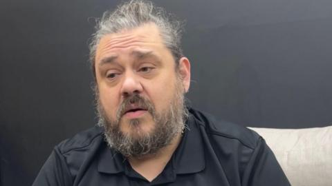 Dan Guilbert a man with black and grey hair with a simimlar coloured beard. He is wearing a black button up shirt and is sitting down. 