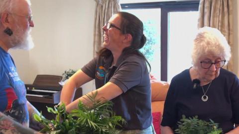 The image show people gathered around a table indoors, taking part in a planting activity. Several potted plants are arranged across the table.