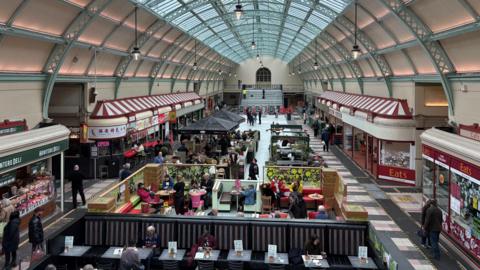The inside of Grainger Market following its renovation. The stalls are lined with red and white tops designed to look like awnings. There are seats and tables for diners and a large space at the far end of the room where events can take place. The roof is arched with ornate ironworks in pale green and a glass ceiling. There are dozens of shoppers and diners scattered about.