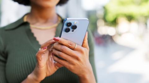 A woman wearing a green top is holding a light blue mobile phone in her hand and using her other hand to press on the screen.