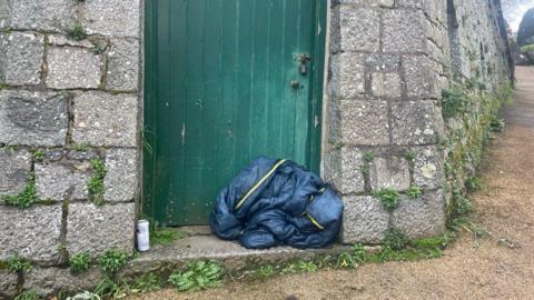 There is a green door in the centre of the photo. It shows a beer can in the doorway and a crumpled up sleeping bag on the right hand side.