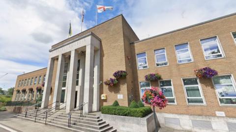 A Google photograph of Rugby Town Hall in Evreux Way. The modern building has steps leading up to the entrance which has pillars and flags flying above it. There are hanging baskets on the front.