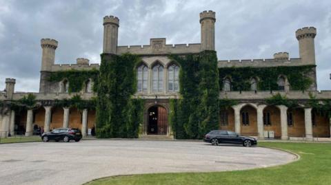 An exterior shot of Lincoln Crown Court which is a Gothic style building with turrets, greenery coming from it and large windows and a wooden front door. Two black cars are parked in front.