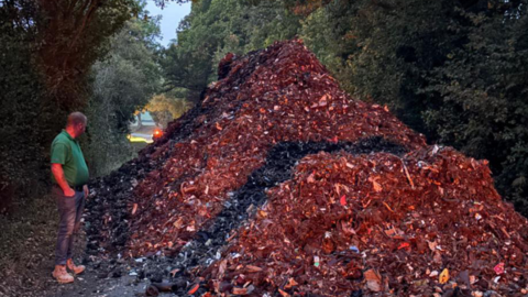A large pile of waste that seems to have been put through a shredder. The waste is blocking a country lane. A man in a green polo shirt stands next to it. The pile is twice his height.