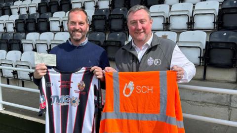 A man wearing a navy jumper holds a cheque and a black and white striped football shirt up, as he stands next to another man, who is wearing a grey gilet with a football emblem on it, over a white shirt. He is holding up an orange hi-vis jacket. Both are standing on a football pitch on a sunny day. Black and white seats are behind them. 