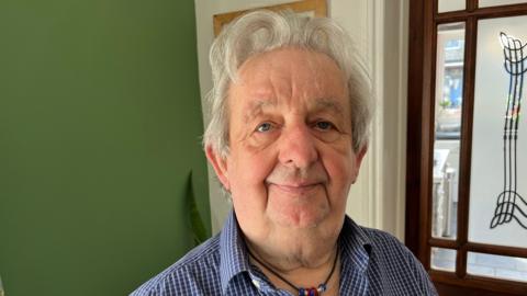 Bill Batrick has grey hair with a slight quiff, he is wearing a blue and white checked shirt, with some beads around his neck.