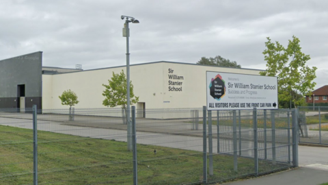 A large cream coloured square building reading "Sir William Stanier School" on the front. It is surrounded by grass and has small trees along a driveway outside. It has a white welcome sign at the front and metal fences.