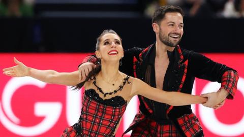 Figure skating pair Lilah Fear and Lewis Gibson perform a routine together, with both dressing in black and tartan outfits, with both smiling