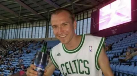A man with short brown hair in a white vest with Celtics in green lettering on it, holding a bottle in a stadium with blue seating and a large number of people sat around him