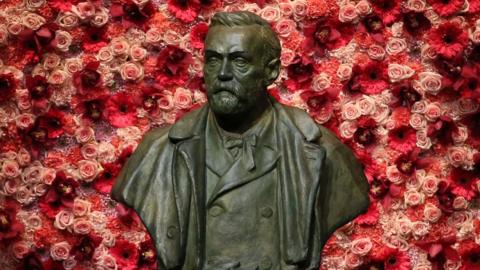 a statue of Alfred Nobel with a background of pink and red roses.