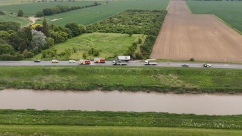 An aerial view of a stretch of road which runs alongside a river. There are several heavy goods vehicles and emergency service vehicles on the road. There is also a lorry which has collided into bushes.