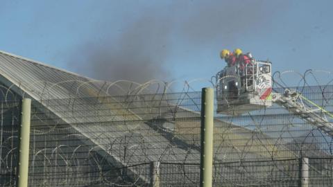 Firefighters in the cage of an aerial ladder platform. There appear to be two of them. They are looking at a roof from which billowing black smoke is coming. The shot has been taken through a barbed wire topped wire fence at Highpoint Prison.