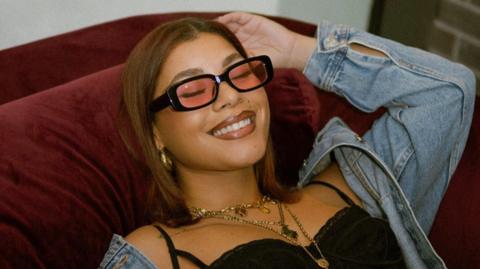 Milena Sanchez pictured reclining on a red velvet sofa. She wears a denim jacket over a black strappy top, styled with red-tinted sunglasses and multiple gold necklaces. She smiles, her eyes closed, as she touches her head with her right hand. 