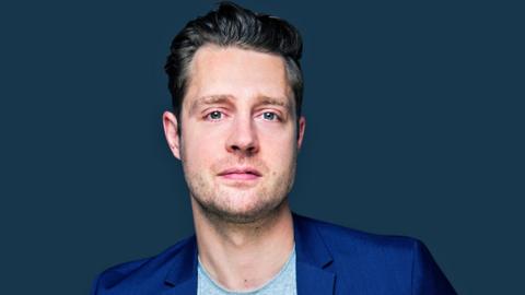 Comedian Max Dickins, wearing a blue suit jacket and light grey t-shirt, fills the frame and looks directly at the camera. He is stood in front of a dark blue background 