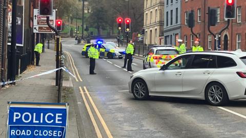 Shot of police vehicles and officers at a crossing on a dual-carriageway. A police sign saying road closed is in the foreground.