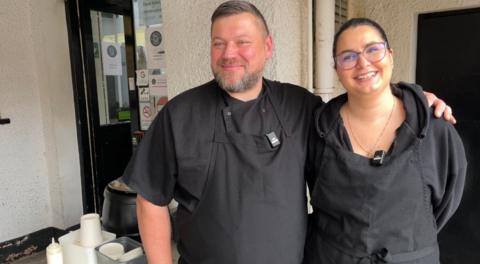 Arek has his arm around Monika - both wearing black clothes and black arpons, smiling facing just off to the right of the shot. You can also see the soup container behind him, as well as a door leading to the kitchen.