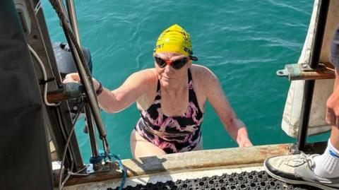 Dot, who wears a swimming costime, cap and goggles, climbs onto a boat from the water.