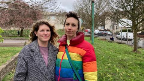 Two women stand together on a patch of grass with bulbs torn out of the grand. On the left, Victoria is wearing a grey coat and on the right Ailie is wearing a rainbow-coloured coat and wooly hat.