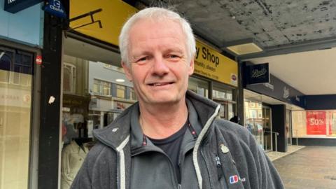 A man standing in front of a selection of high street shops wearing a grey fleece jacket. 