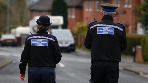 Two police community support officers wearing a black uniform with blue and white chequered detailing on the shoulders, are walking away from the photographer. On the left is a female officer wearing a bowler-style police hat and on the right is a male officer wearing a flat-style police hat. They appear to be walking down the centre of a residential street.