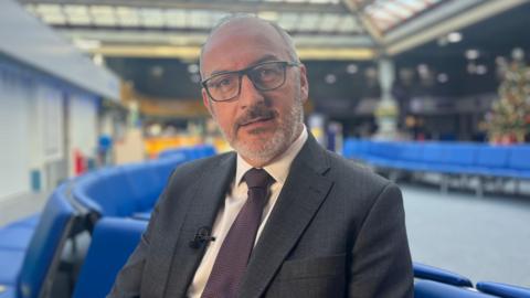 Christophe Mathieu - a man wearing black rectangular glasses, a grey suit with a white shirt and purple tie, sitting on blue chairs in the harbour ferry terminal.