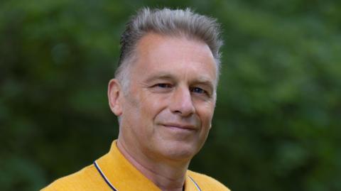 A man with short grey hair smiles at the camera as he stands outside. He wears a yellow polo shirt. There are green bushes and trees behind him.