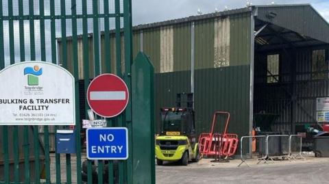 Newton Abbot waste transfer station. A green building with a green gate in front of it. There are bins inside and in front of the building. There is a no entry sign on the gate. There is also a Teignbridge Council sign on the gate.