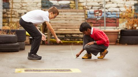 A pupil draws a line on the playground using chalk while another points