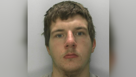 A mugshot of a 19-year-old with brown hair, staring blankly at the camera. He has spots on his chin. He has a grey sweatshirt on and is stood in front of a grey background.