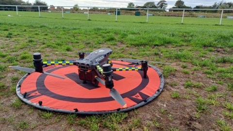 A police drone stationery in a field. The machine is black and connected to a circular orange structure.