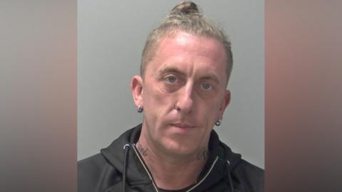 A police custody image of a man with a blond top knot, a black hoodie and small silver hoop earrings. He has some tattoos on his neck.