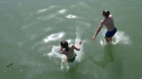 Two children jump into a body of water wearing swimming costumes