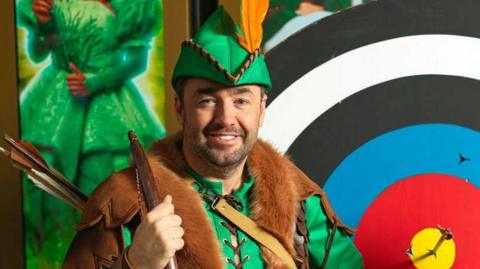 Jason Manford in a green Robin Hood pantomime costume, with a green peaked hat with a yellow feather, a green laced up jumper and a brown fur shrug, holds a wooden bow and wears a quiver of arrows in front of an archery target, which has black, white, blue and red rings around a yellow bullseye