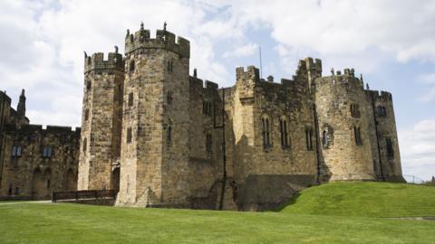 An exterior view of Alnwick Castle in Northumberland. A grey brick, turreted castle with long windows and archways