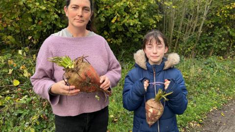 Dawn Qince, with brown hair and wearing a purple jumper, is holding a Mangelwurzel to carve and and her daughter Cherry, wearing a blue coat, is holding a carved lantern. They are both stood outside with bushes in the background.