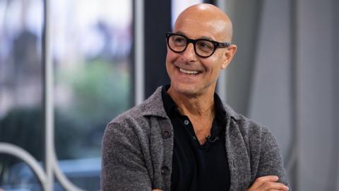 Stanley Tucci smiles and looks away from camera with his arms folded, taken on the set of the Today show in 2023