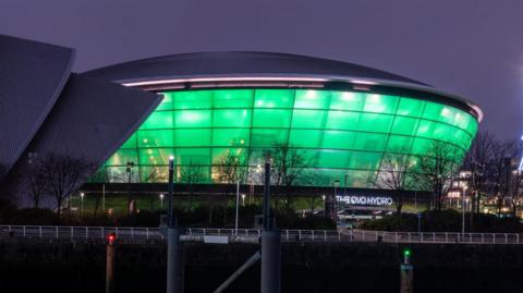 A large events arena with its  windows lit up in green in front of a river