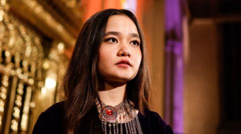 Nila Ibrahimi stands in front of a gold and purple background, wearing a dark top and a necklace with red colouring