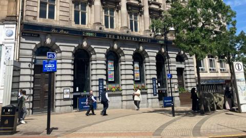 People walk by the Banker's Draft pub on a sunny day. The former bank building has grey columns and ornate detailing.