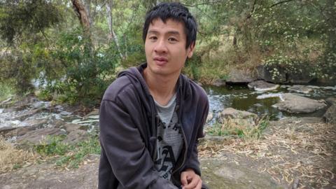 Tim Chan is sitting next to a stream in an outdoor area. There are trees visible behind him, and he is wearing a dark grey hooded sweatshirt and looking straight at the camera. 