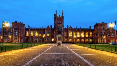 The Queens University in Belfast at twilight.  Several lights are on inside the large, red brick building, which is approached by a cobbled path flanked by neatly mowed lawns. 
