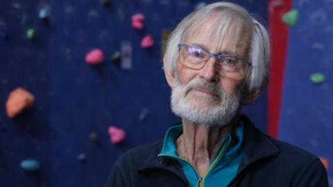 Martin Boysen is sitting in front of an indoor climbing wall. He is wearing glasses and has grey hair and a beard. He is wearing a blue sports top and a navy jacket. 