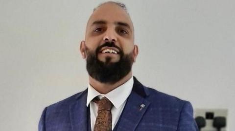 A smiling man with a black beard and a balding head. He is dressed in a navy suite with a white shirt and brown tie. 